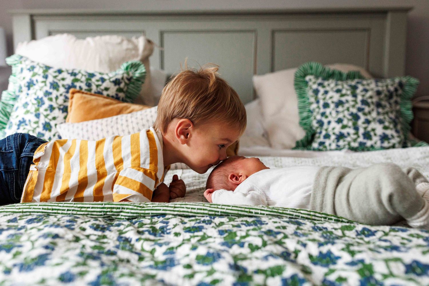 Toddler giving a gentle kiss to a newborn baby while lying together on a bed during a lifestyle newborn photography session.