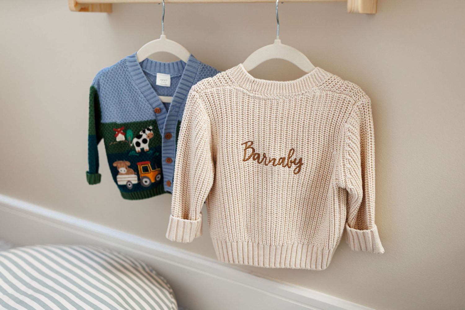 Two baby cardigans on white hangers, a cream knit sweater embroidered with Barnaby and a blue and green one with farm animal appliqués.
