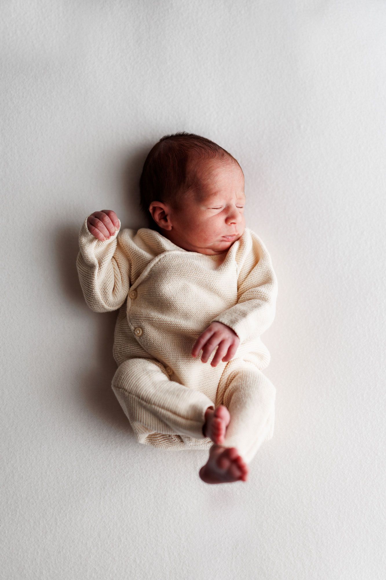 Sleeping newborn baby in a cream knit outfit lies on a soft white blanket, tiny hands curled and bare feet showing.