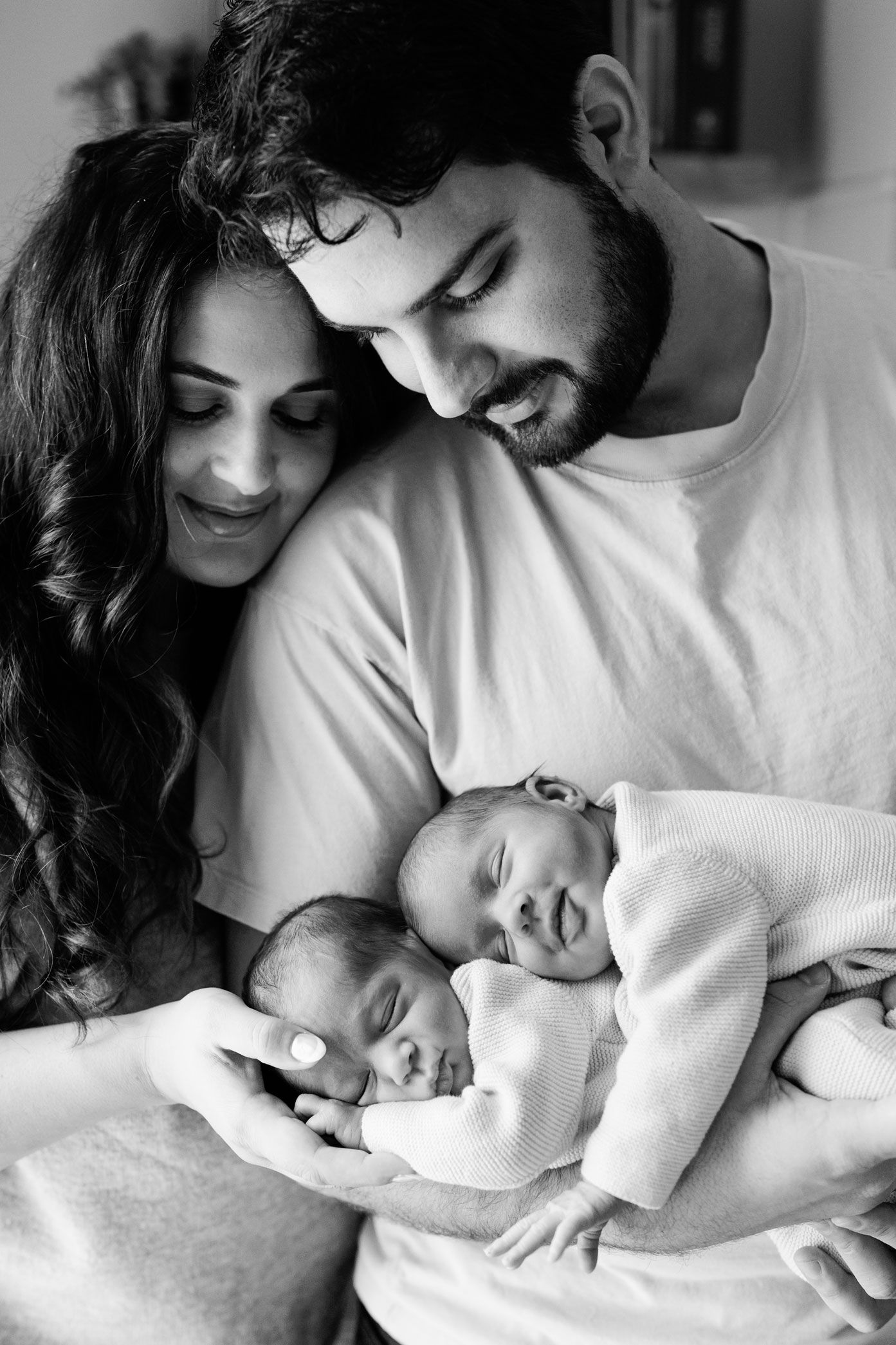 Black and white portrait of smiling parents gazing lovingly at their two sleeping newborn twins cradled in the father's arms.