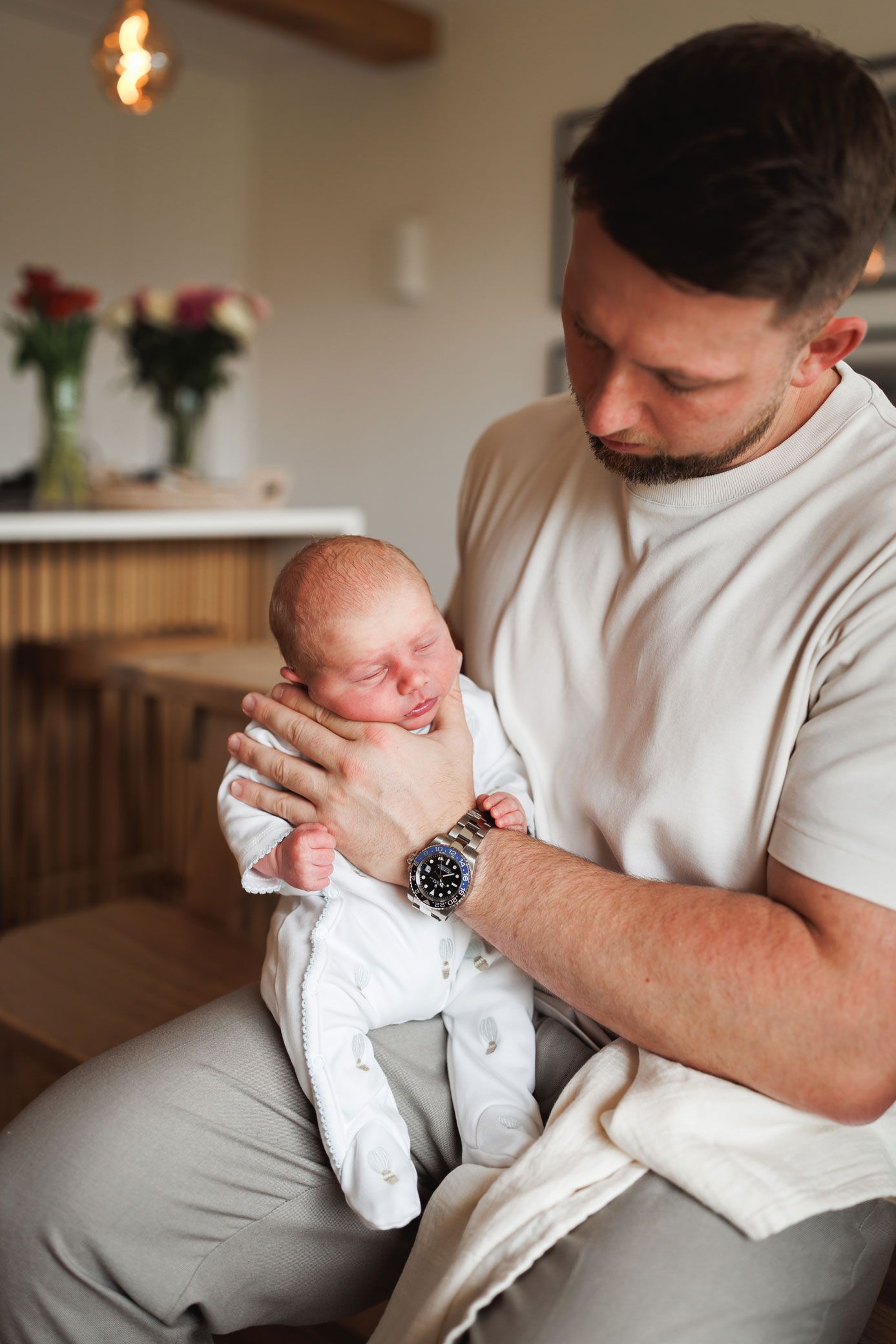 A father in a cream t-shirt gently cradles his sleeping newborn baby in a white outfit in a bright, cozy kitchen with fresh flowers.