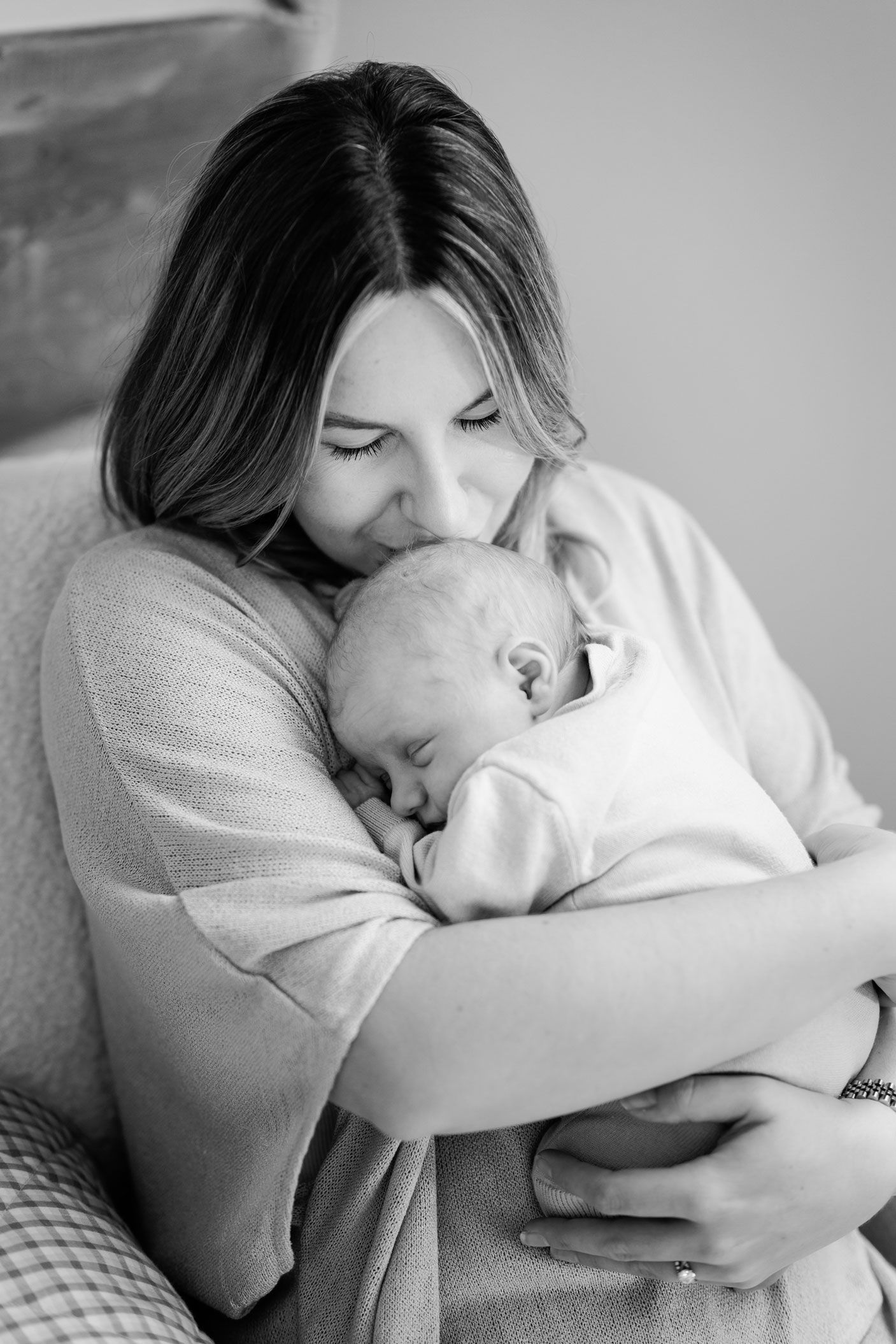 Black and white photo of a smiling mother with long hair tenderly cradling and kissing her sleeping newborn baby in a cozy home setting.