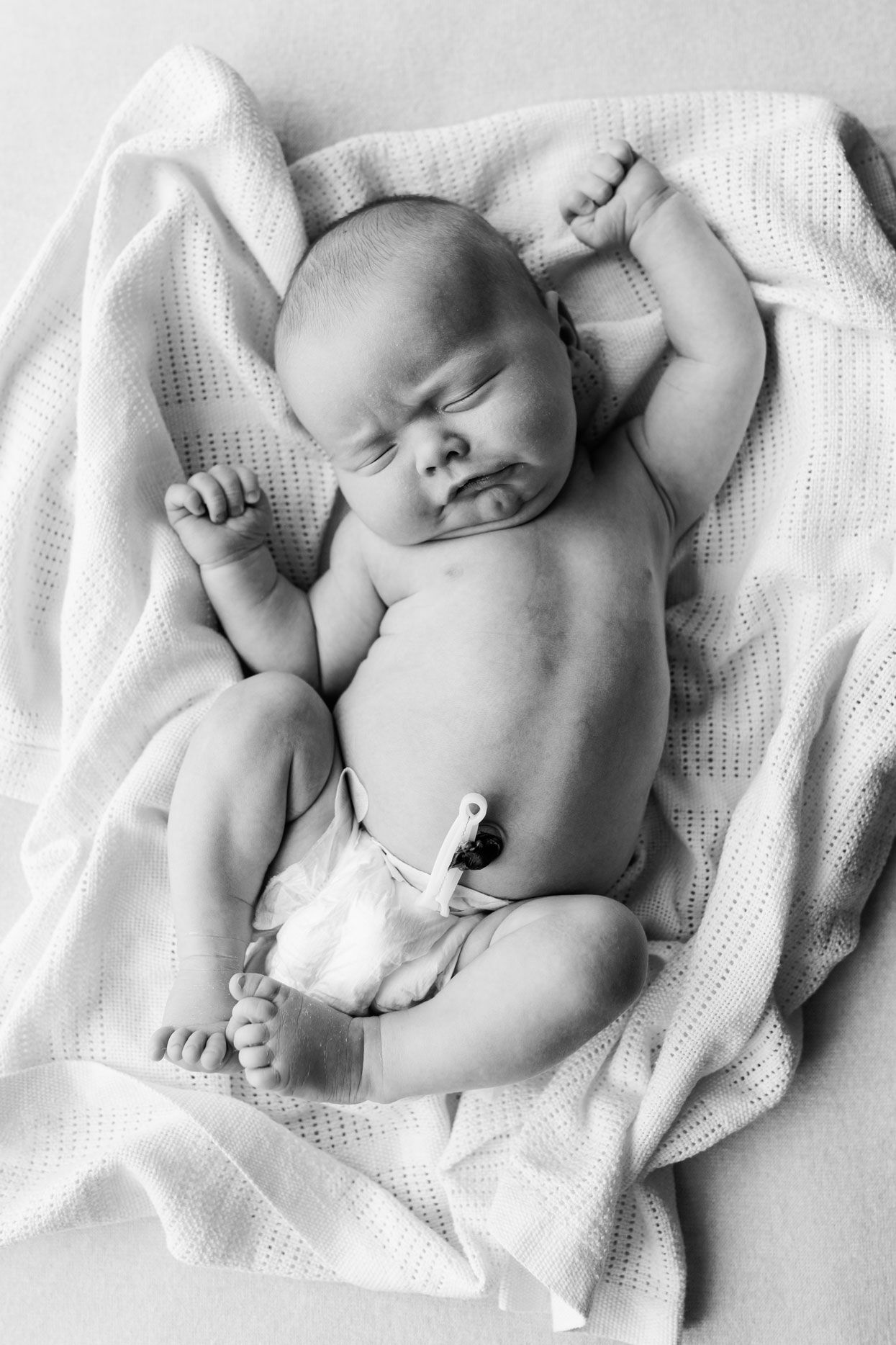 Black and white photo of a newborn baby lying on a soft white blanket, arms raised, wearing a diaper with umbilical cord clip.