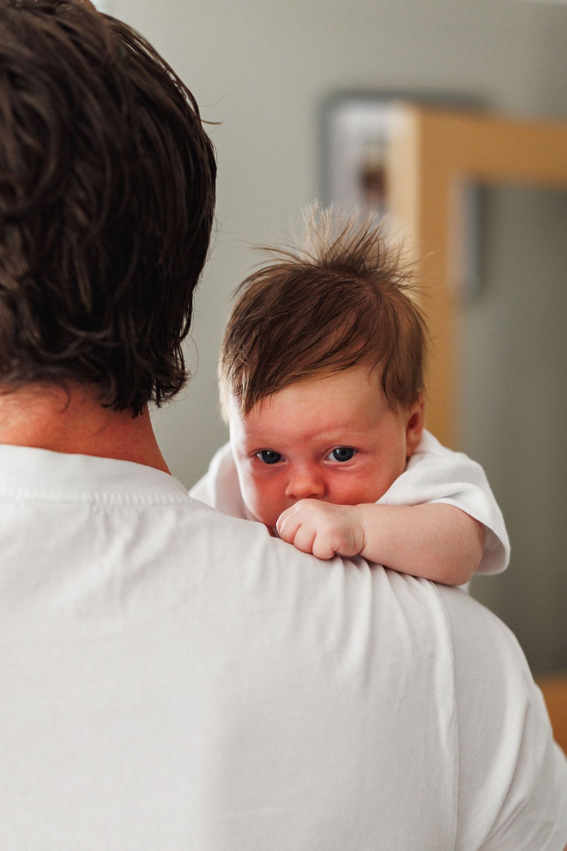 A newborn baby with spiky brown hair rests on an adult's shoulder, wearing a white outfit indoors.