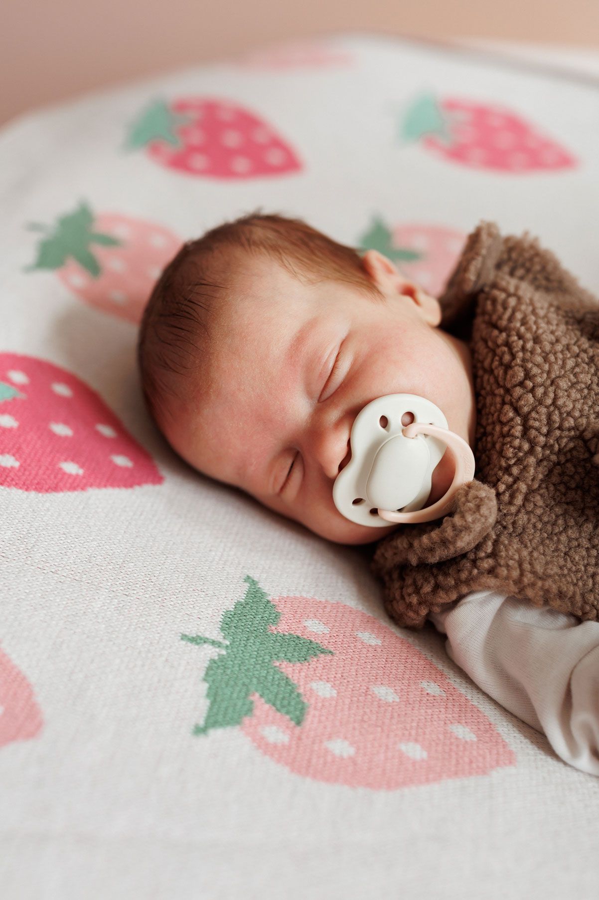 Sleeping newborn baby with a white pacifier wearing a brown fleece sweater, resting on a soft blanket with pink strawberry print.