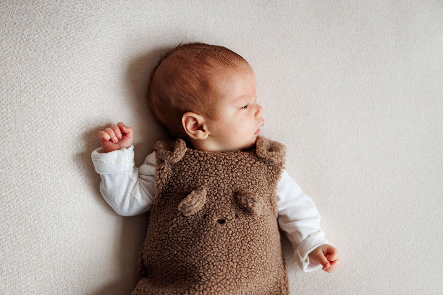 Newborn baby sleeping on a soft cream blanket, wearing a brown teddy bear fleece vest over a white long-sleeve onesie.