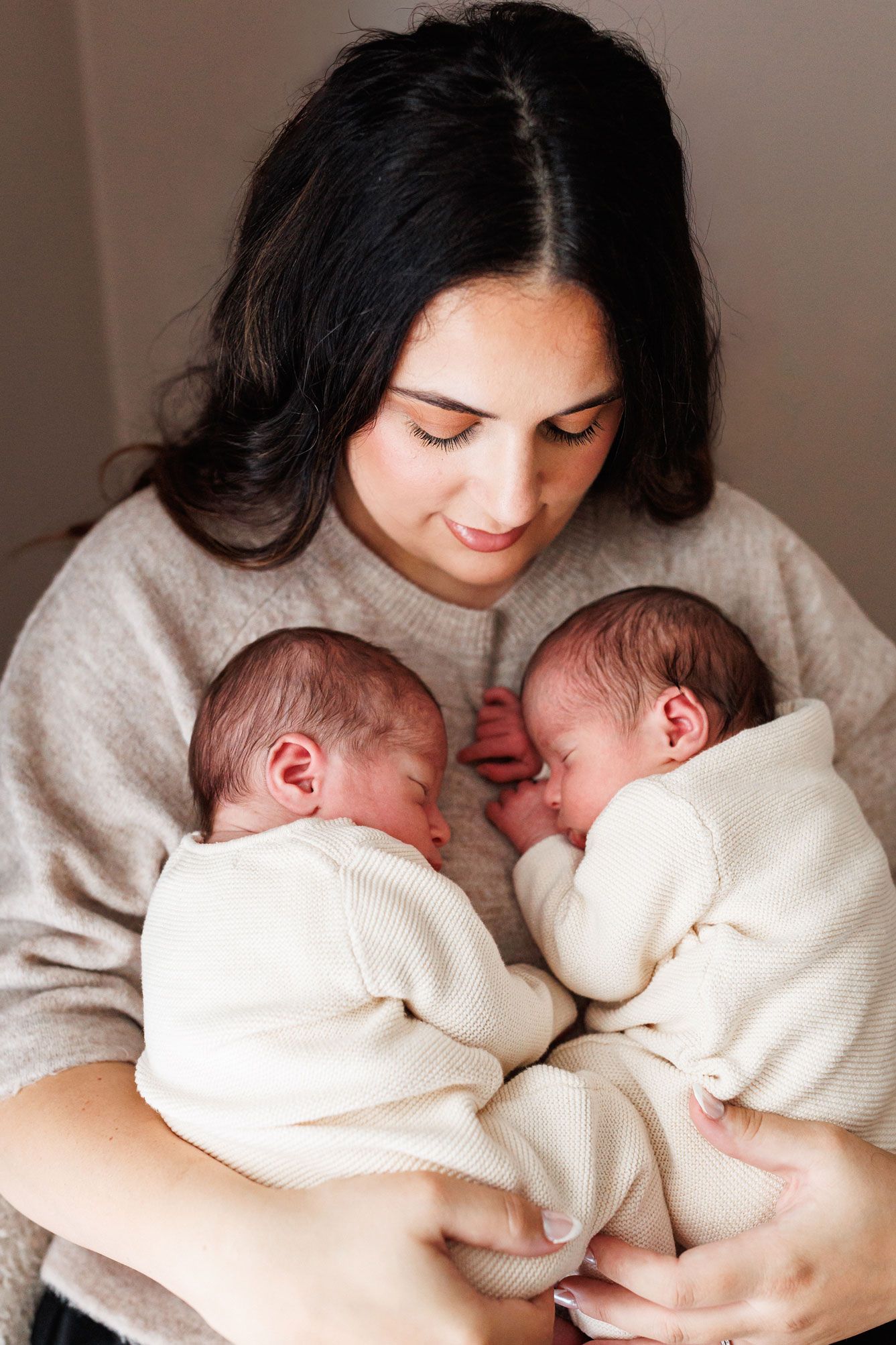 Smiling mother in a cozy beige sweater holds her two sleeping newborn twin babies wrapped in cream outfits against her chest.