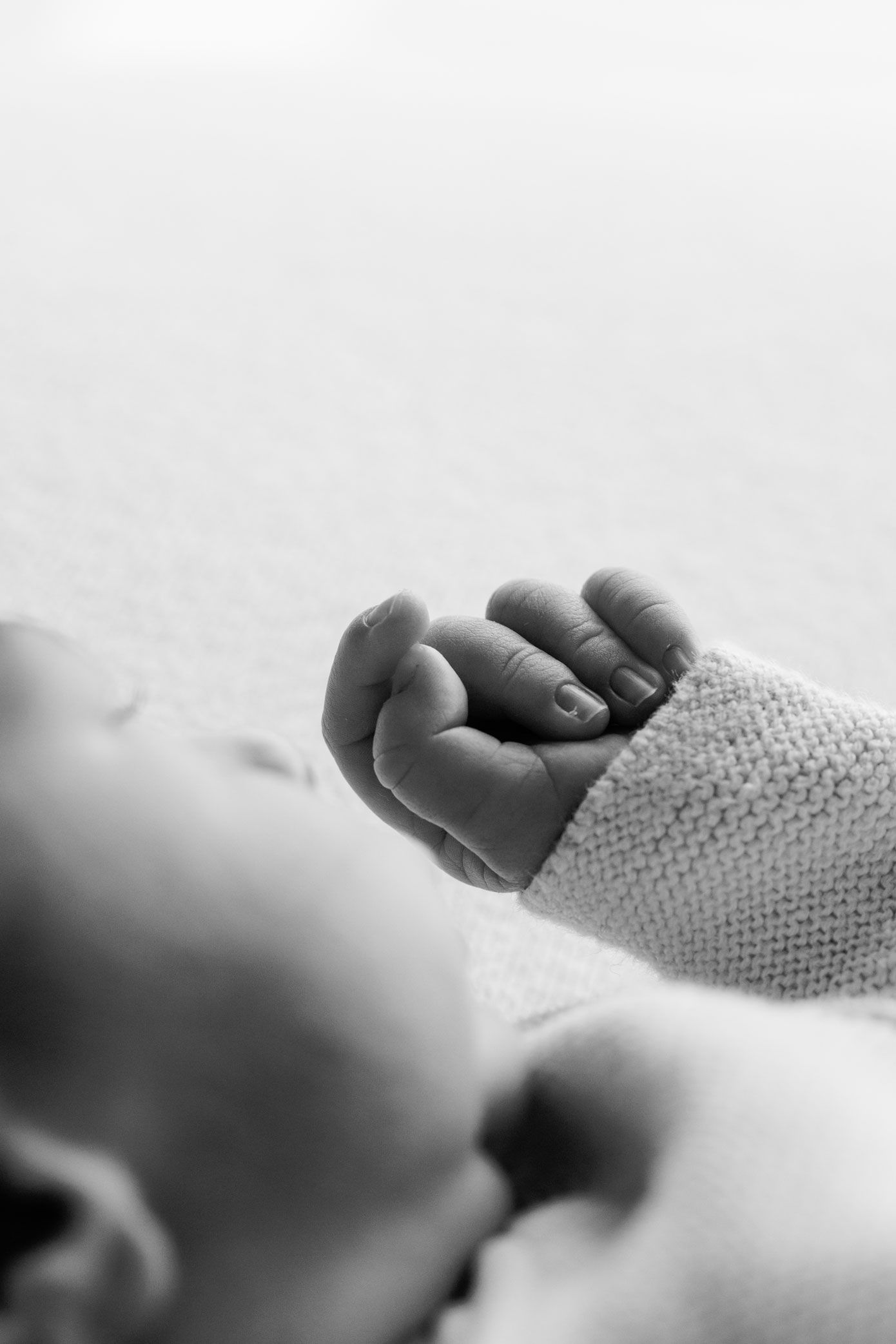 Black and white close-up of a newborn baby's tiny curled fist resting on a soft blanket, wearing a knitted sweater.