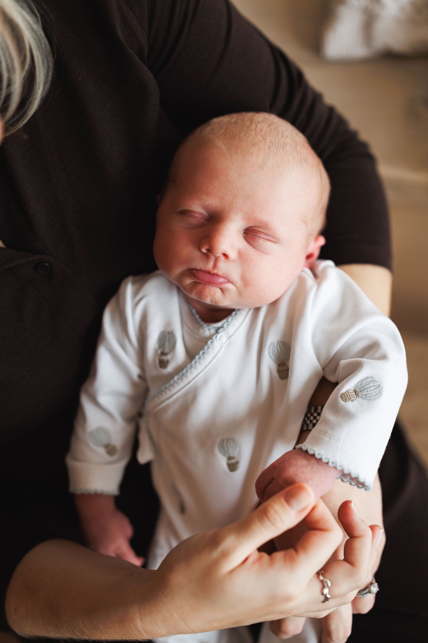 Sleeping newborn baby in a white onesie with hot air balloon print being held in the arms of an adult wearing rings.