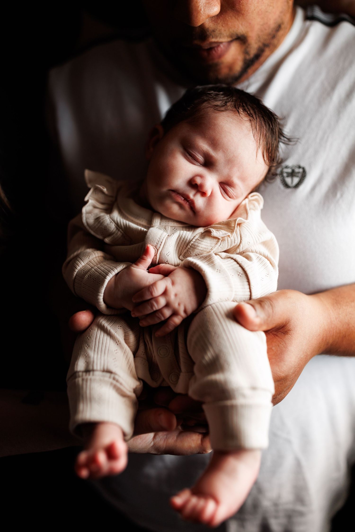Father gently cradling sleeping newborn baby dressed in cream striped onesie, tender moment captured in warm light.