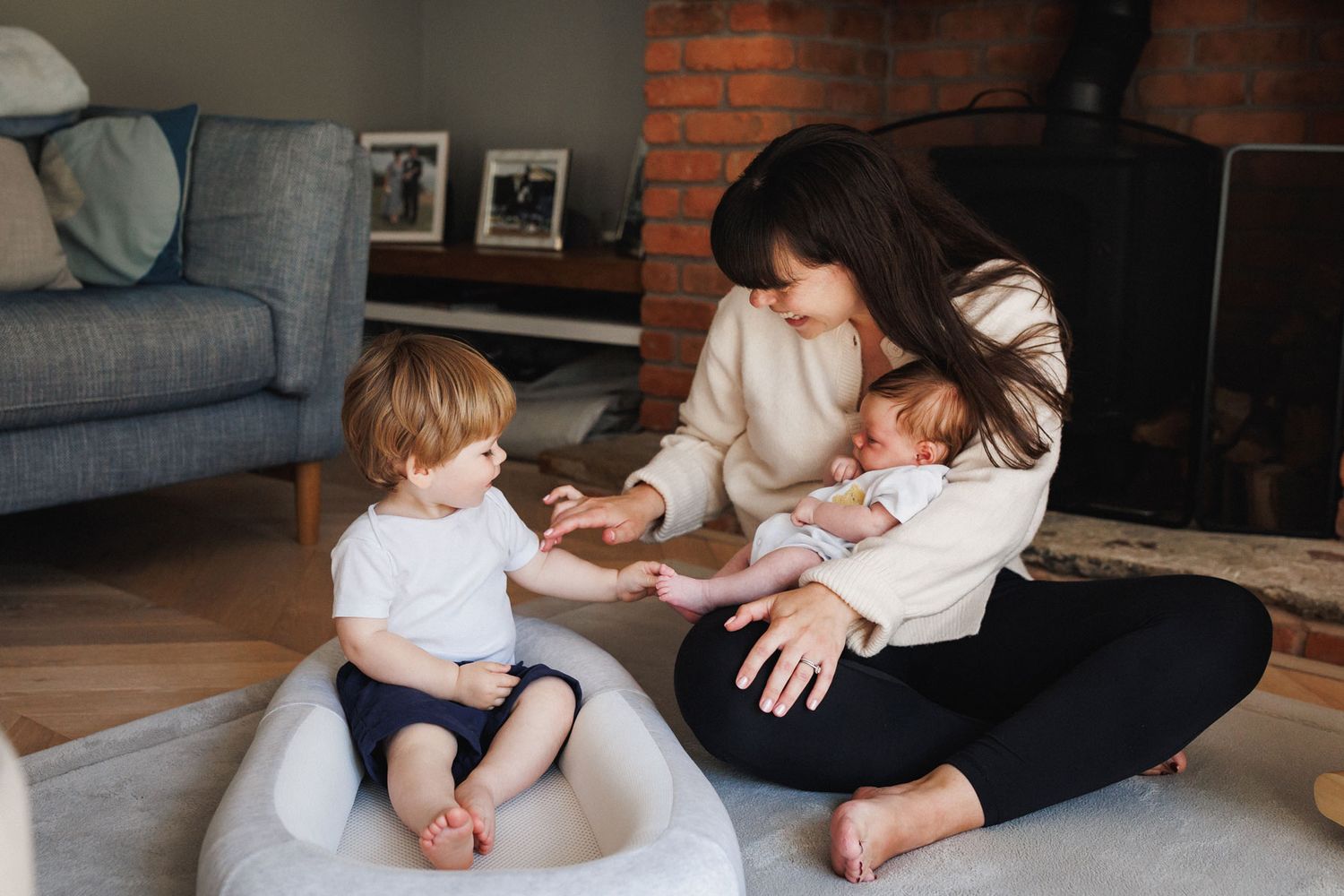 Mother sitting on floor holding newborn baby while interacting with toddler son in cozy living room.