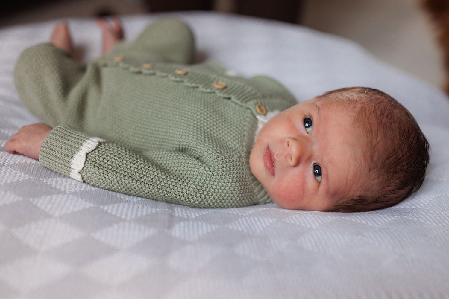 Newborn baby in sage green knit romper lying on white surface.