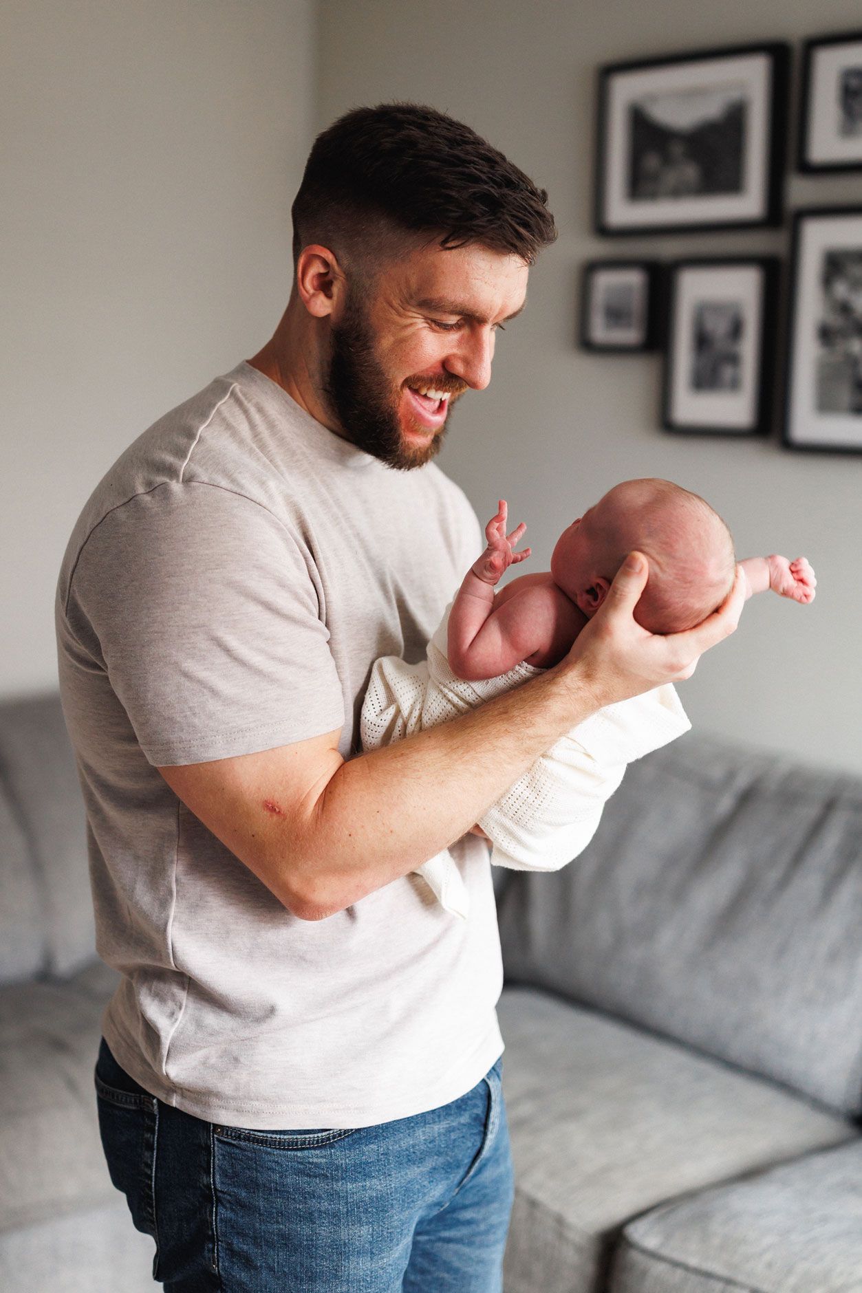 Smiling bearded man holding a newborn baby in his arms while standing in a modern living room.