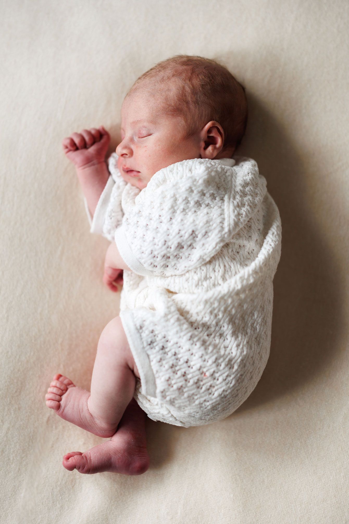 Peaceful newborn baby sleeping in a white knit romper on a soft cream blanket.