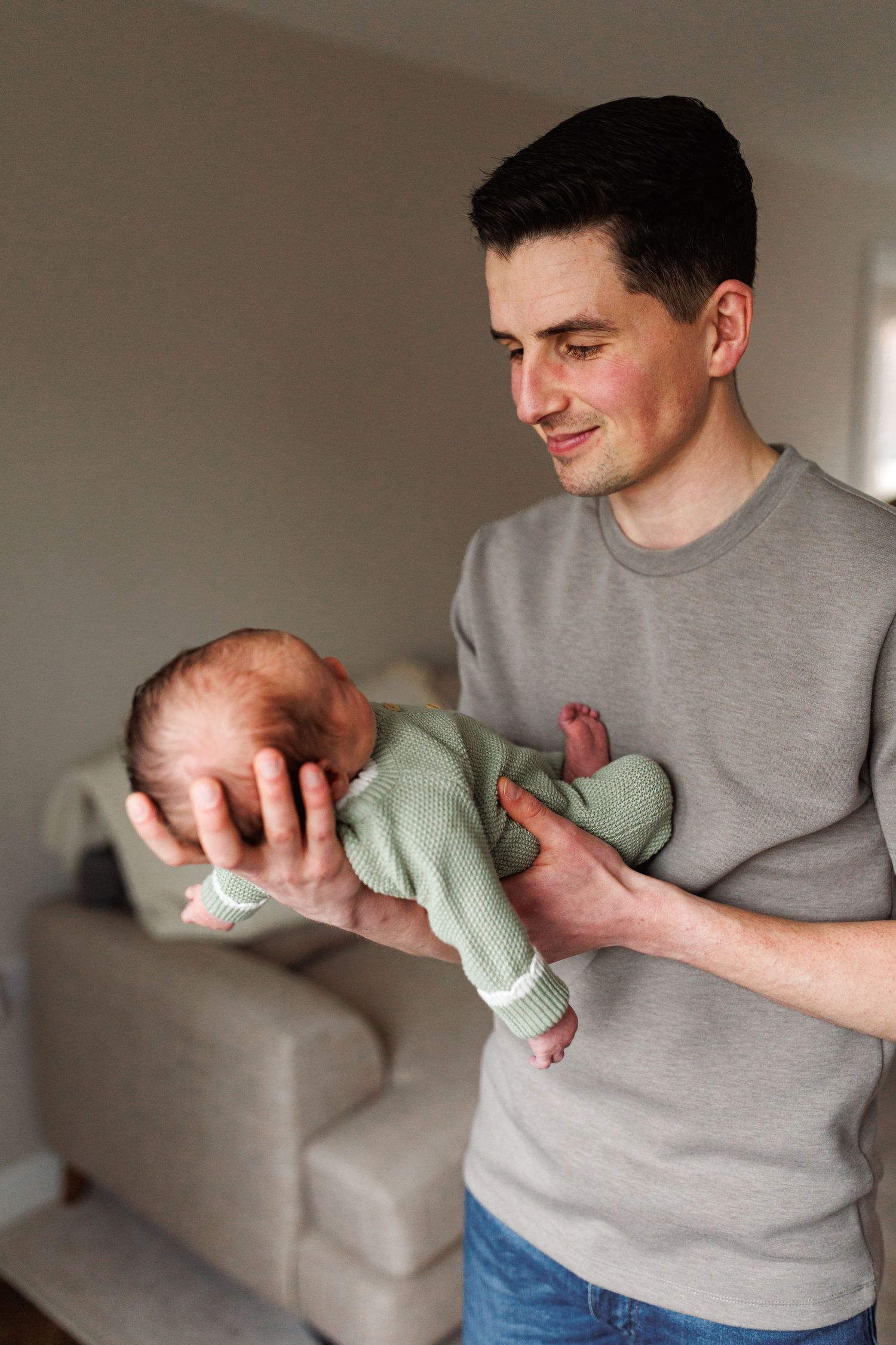 A smiling father in a gray shirt cradles his newborn baby dressed in a green onesie.