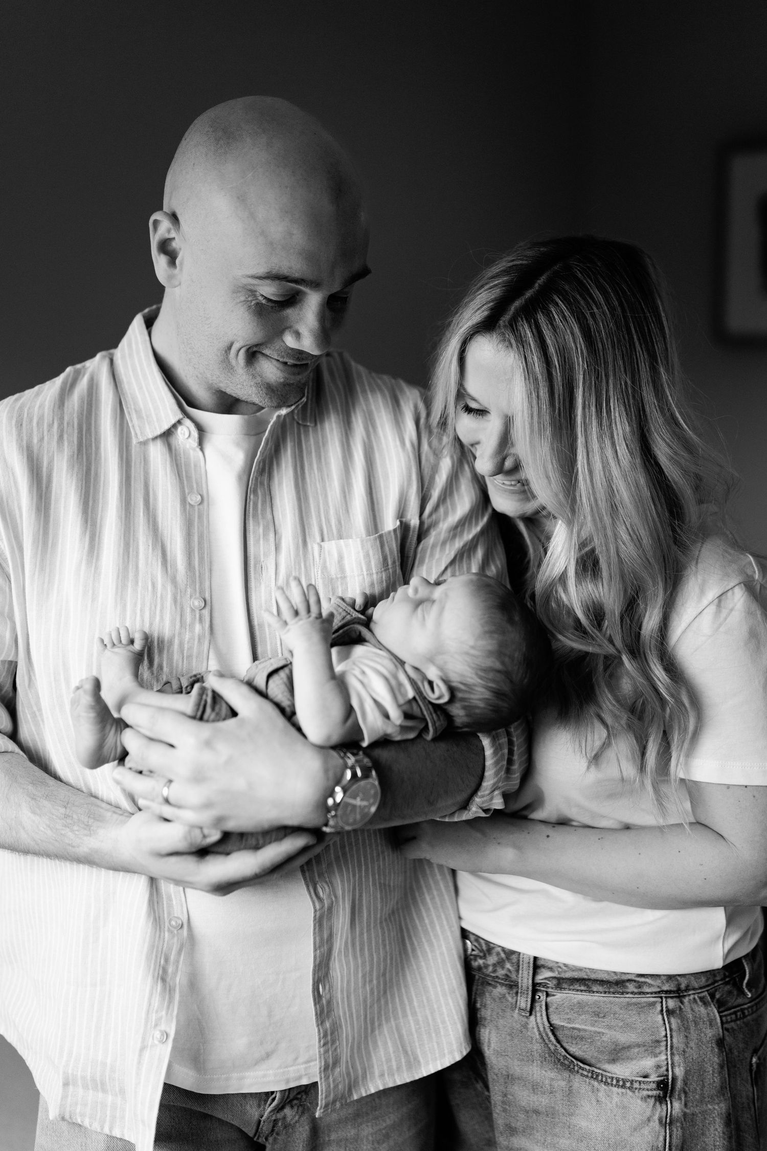 Black and white photo of parents lovingly cradling their newborn baby together indoors.