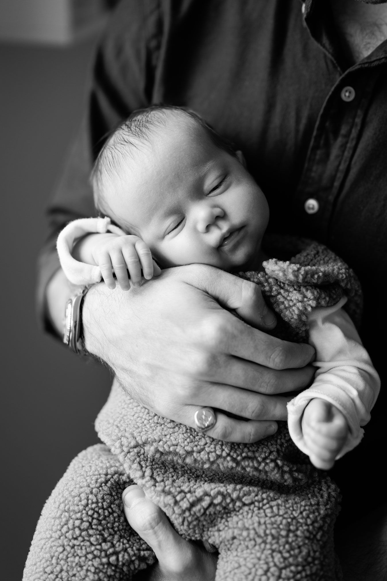 Black and white photo of a sleeping newborn in a fuzzy sherpa outfit, cradled gently in a parent's hands.