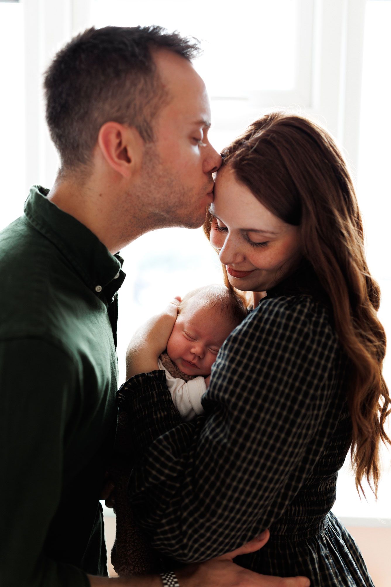 A father kisses the forehead of a smiling mother as she cradles their sleeping newborn baby in a bright, softly lit room.