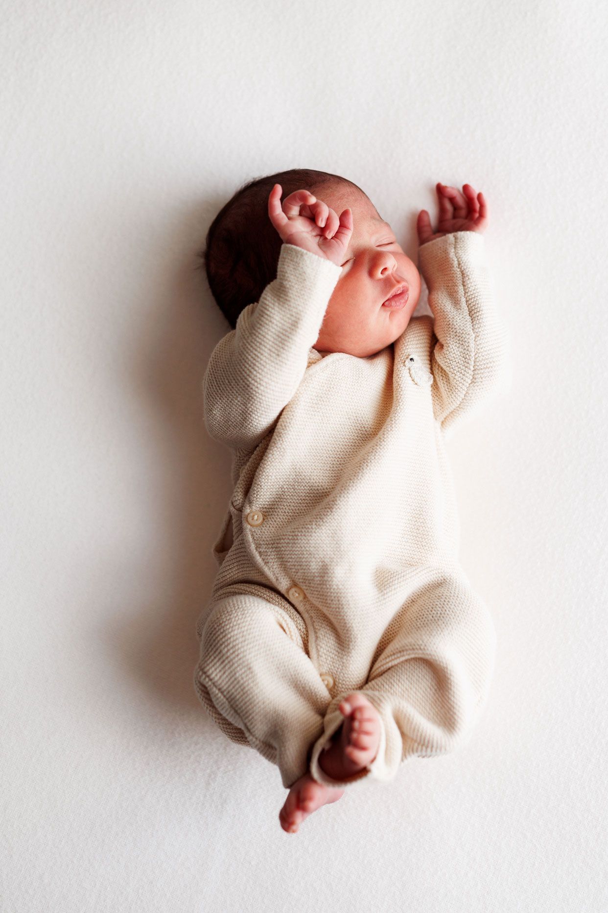 Sleeping newborn baby in a cream knit button romper stretches both arms overhead on a soft white blanket.