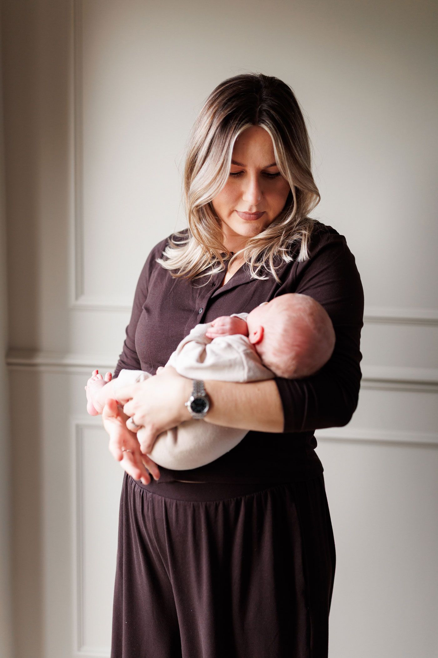 Woman with blonde hair in a dark brown outfit gently cradles a sleeping newborn baby in her arms against a soft neutral wall.