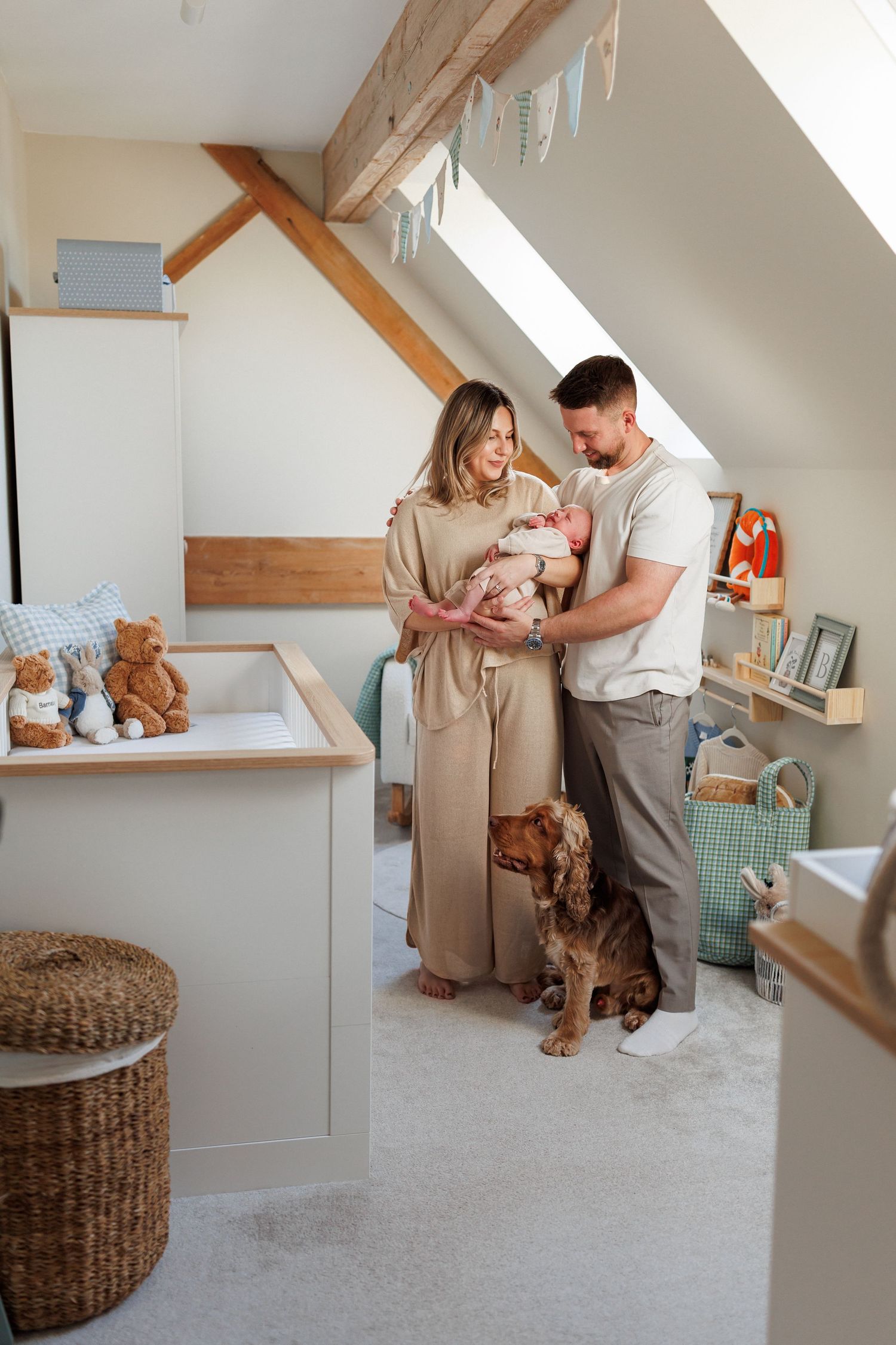 Parents holding their newborn baby beside a crib with teddy bears in a bright attic nursery with a golden retriever at their feet.