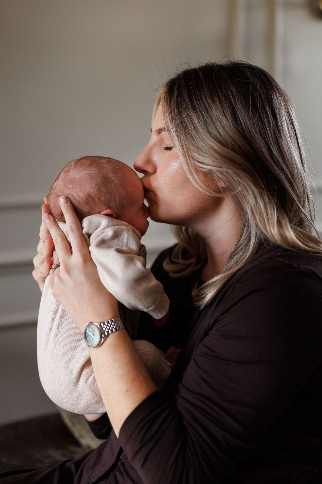 A woman with long blonde hair and a watch gently kisses the head of a newborn baby wrapped in a cream outfit.