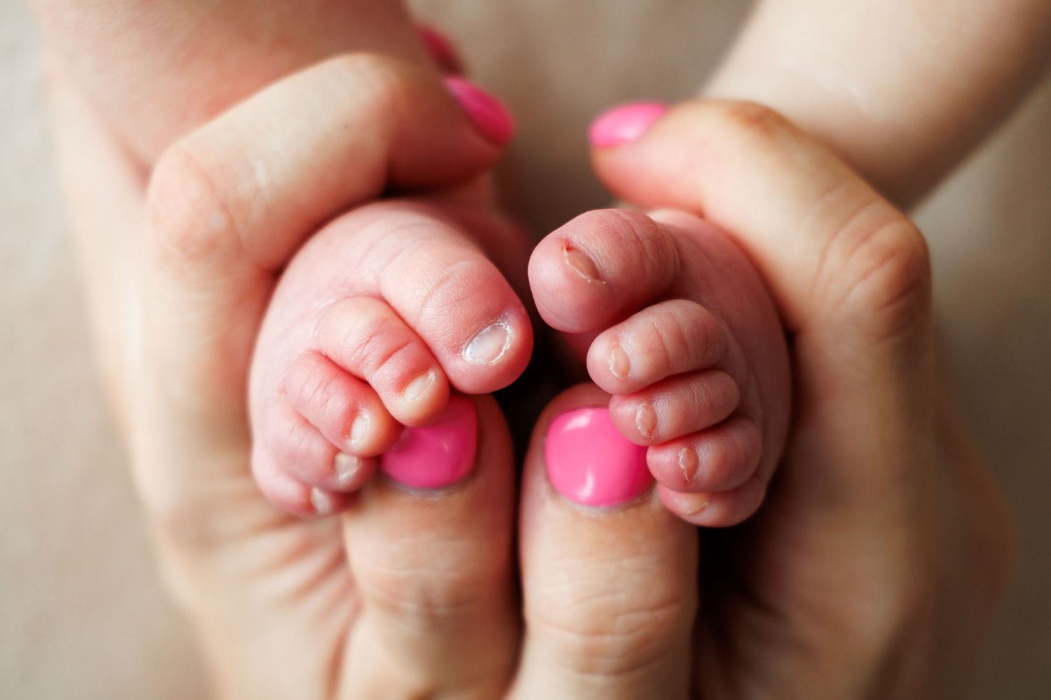 Mother with pink nails gently cups her newborn baby's tiny feet in her hands.