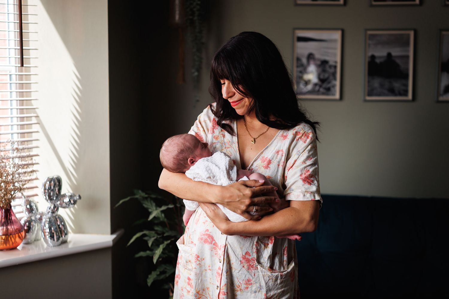 Dark-haired mother in floral robe tenderly holding her newborn baby in sunlit room.