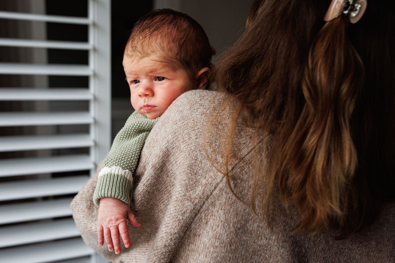 A mother in a beige sweater holds her newborn baby over her shoulder near a window with shutters.