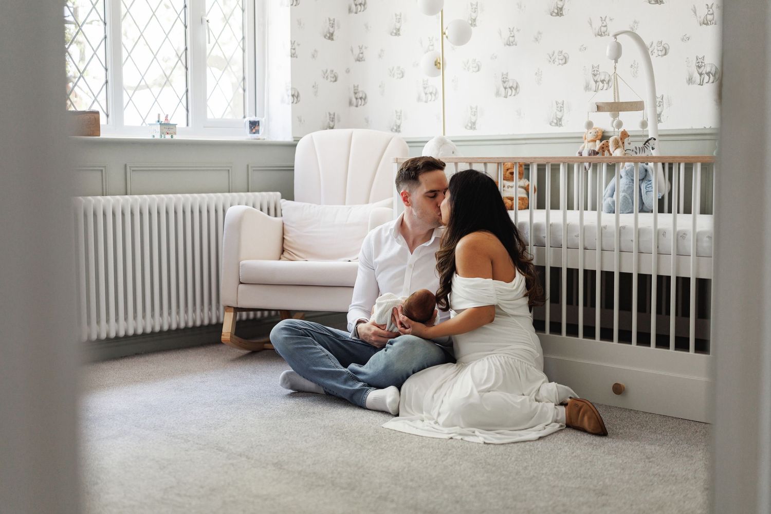 Young couple sitting on nursery floor kissing while holding their newborn baby near a white crib.
