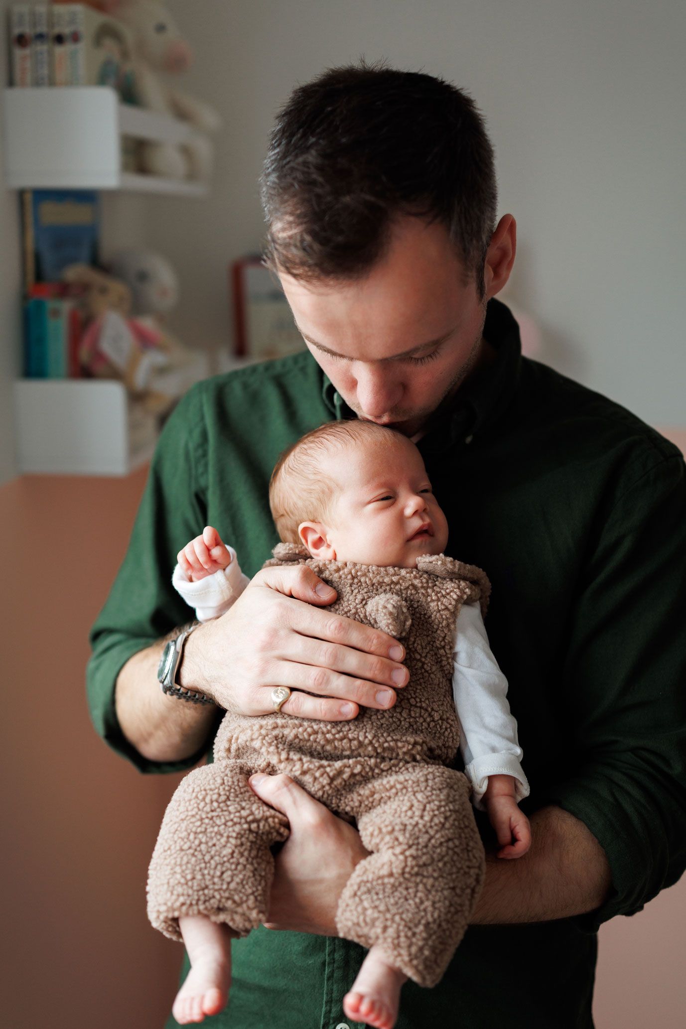 A father in a green shirt gently kisses the head of his sleeping newborn, who wears a cozy brown fleece romper, in a softly lit nursery.