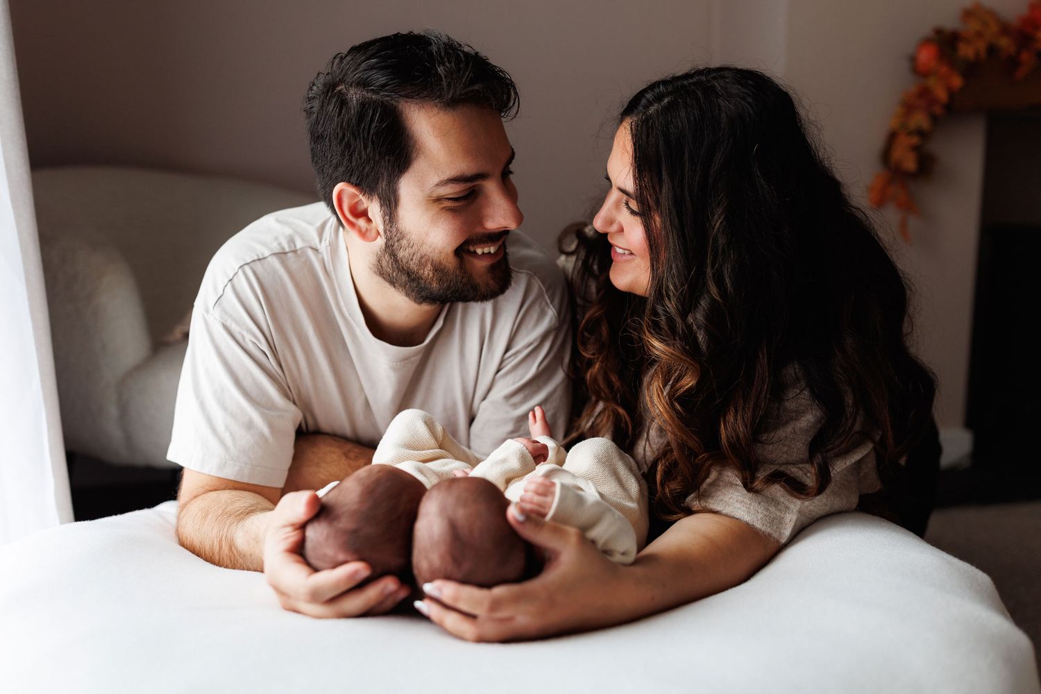 Smiling couple lying on a white bed gazing at each other while gently holding their newborn baby's tiny feet during a newborn photo session.