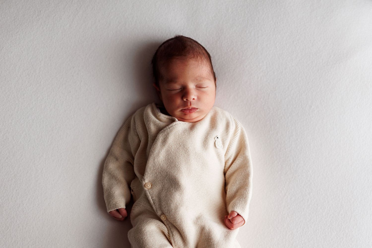 Sleeping newborn baby in a cream knit wrap-style outfit lying peacefully on a soft white blanket.