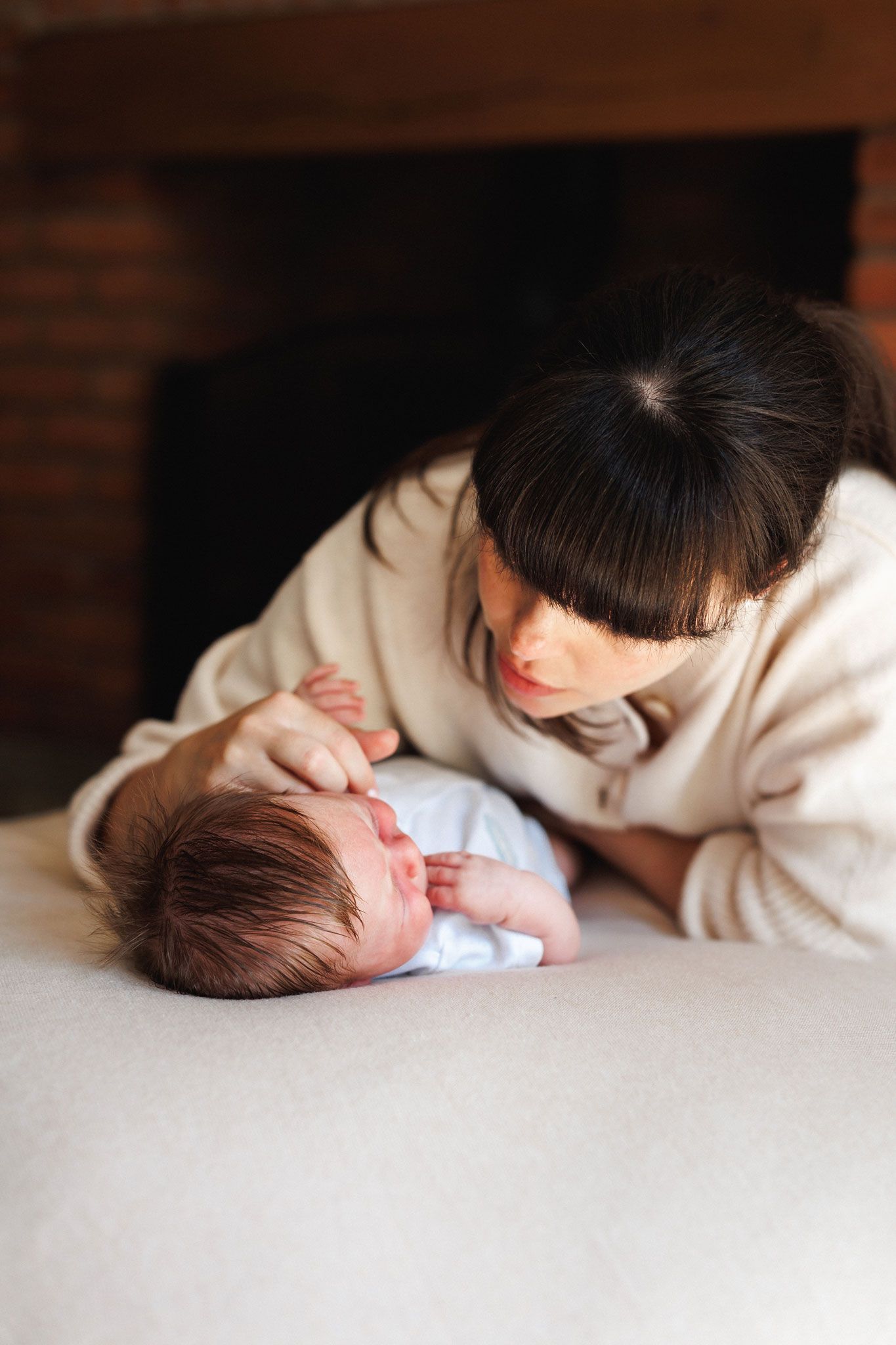 A dark-haired mother in a cream sweater gazing lovingly at her newborn baby on a sofa.