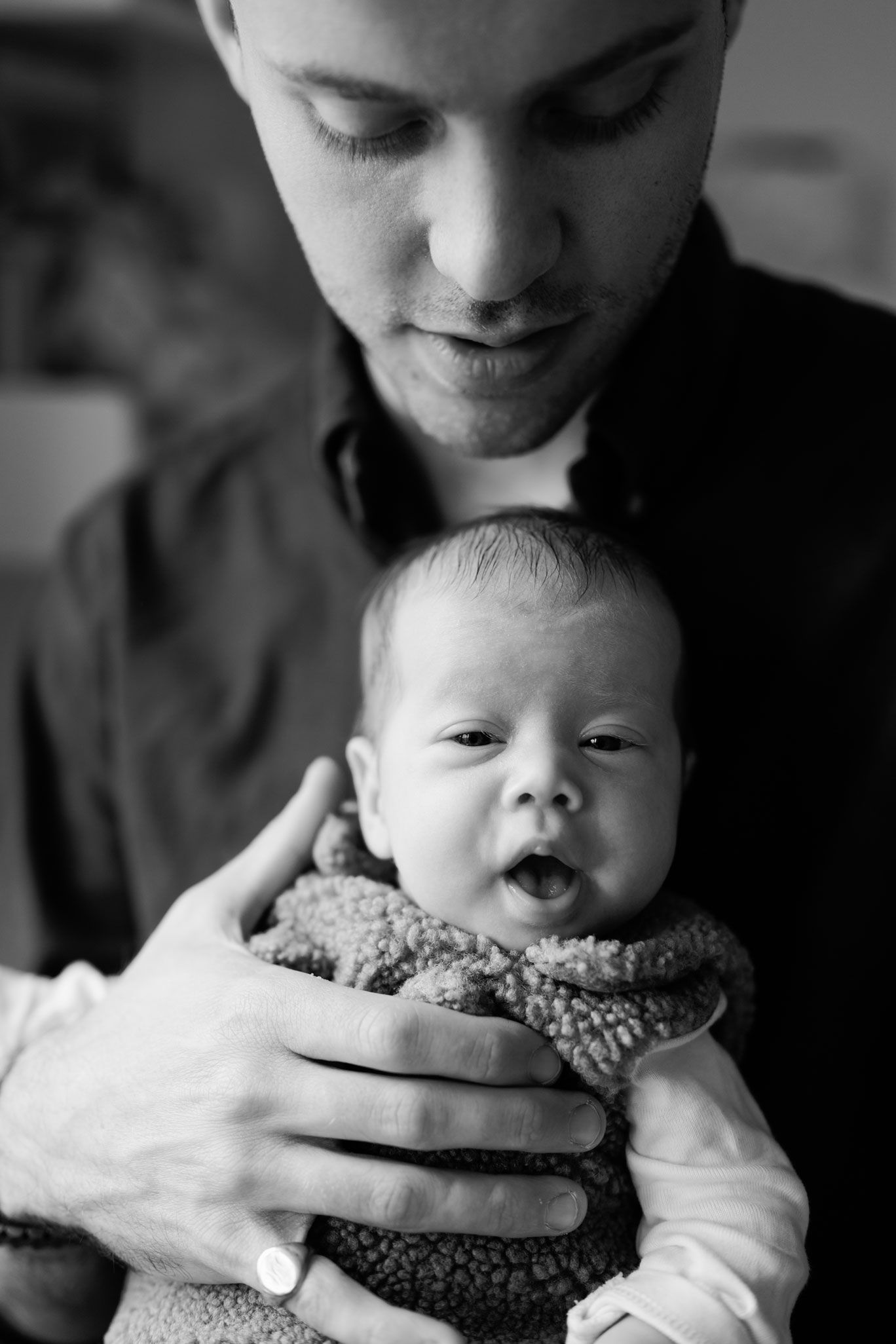 Black and white photo of a father gently cradling a wide-eyed newborn baby in a cozy fleece wrap against his chest.