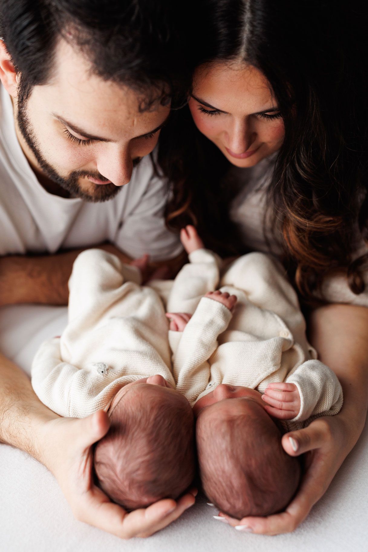 Smiling parents gaze down at their two newborn twins dressed in cream outfits and lying side by side on a soft white blanket.
