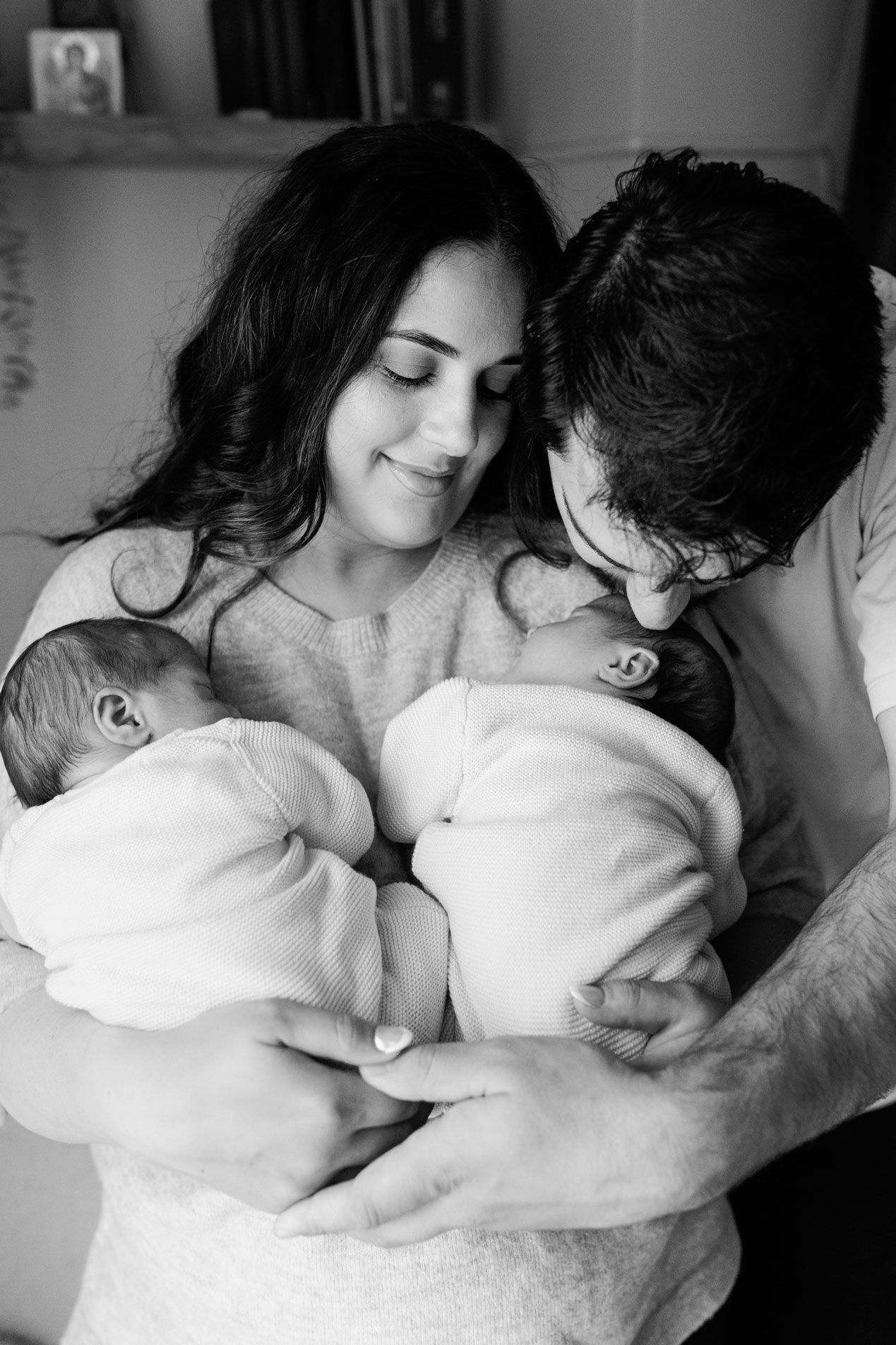 Black and white family portrait of smiling parents tenderly cradling their newborn twins wrapped in white as the father kisses one baby.