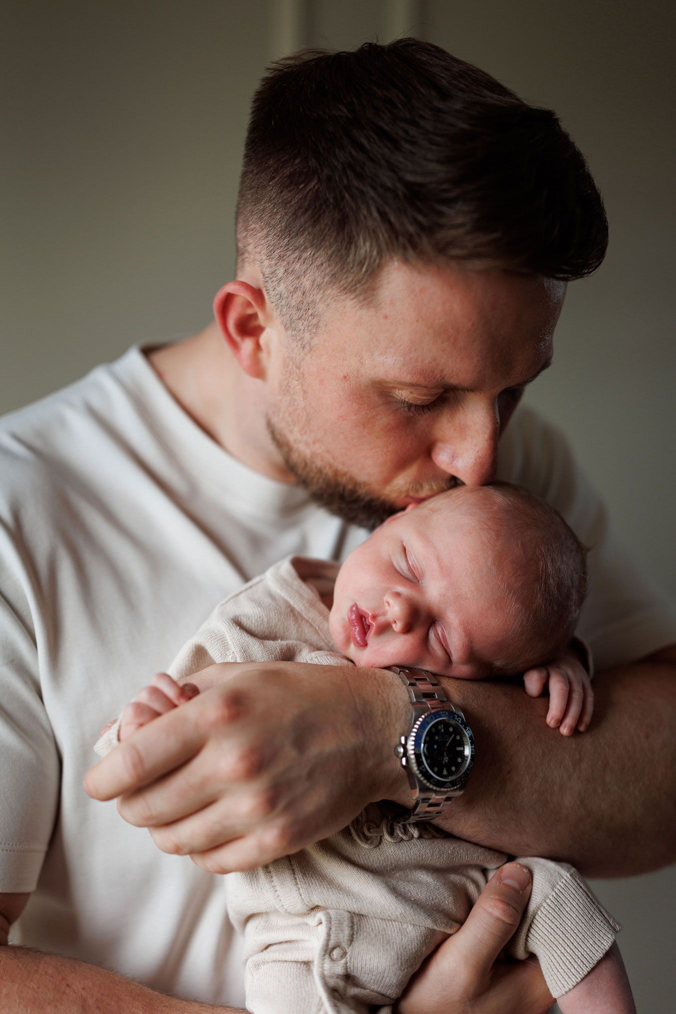 A bearded man in a white t-shirt and wristwatch tenderly kisses the forehead of a sleeping newborn baby wrapped in a cream outfit.