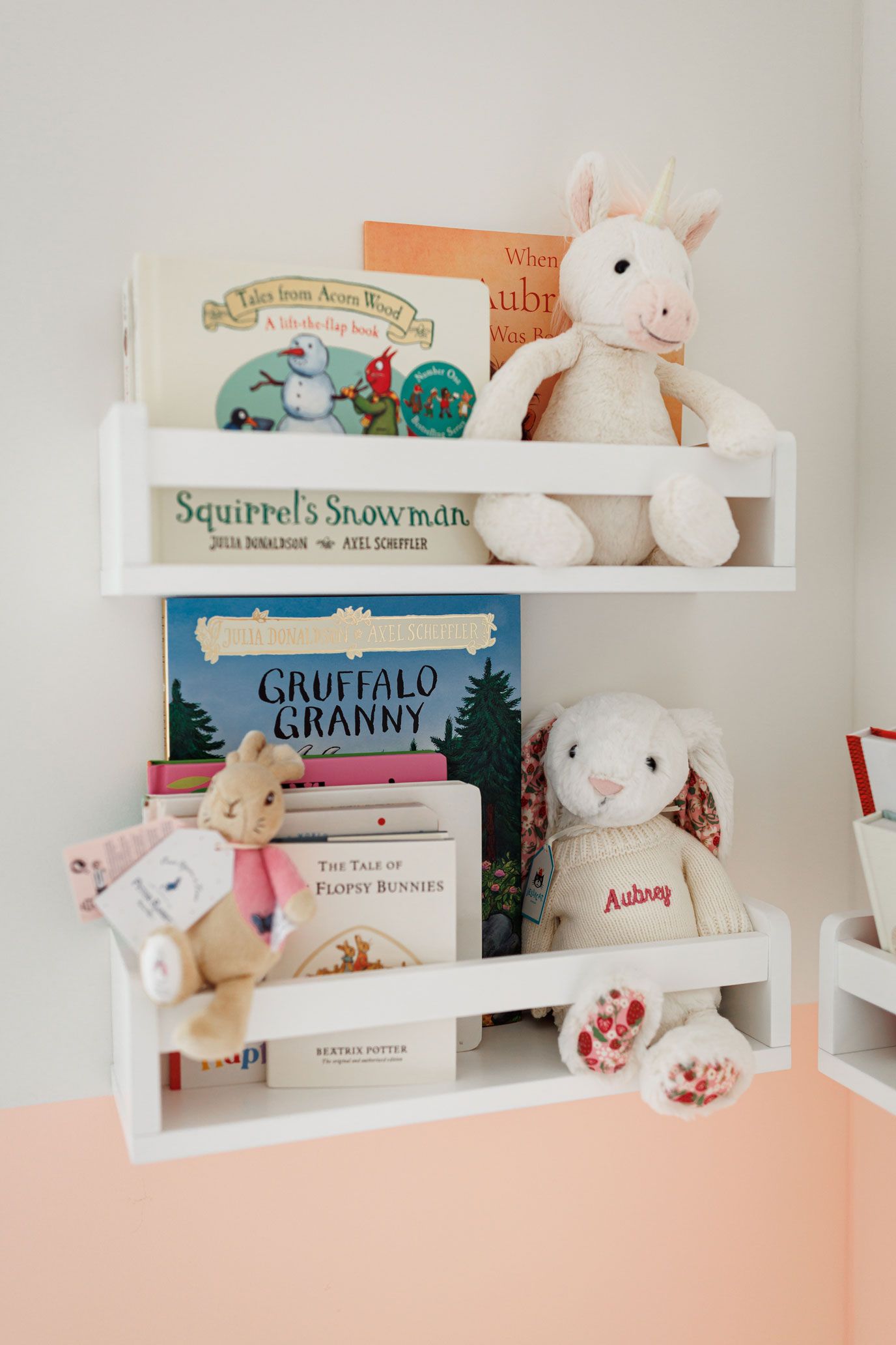 White nursery wall shelves holding children's books like Gruffalo Granny and Squirrel's Snowman, with plush bunny toys against a peach wall.