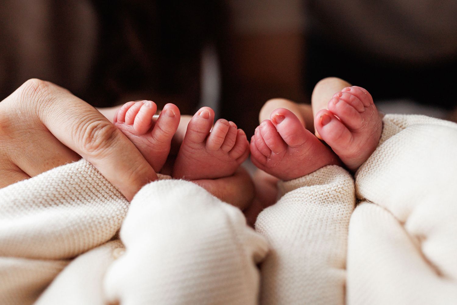 Two tiny newborn baby feet side by side, gently held by a parent's hand and resting on a soft cream knit blanket.