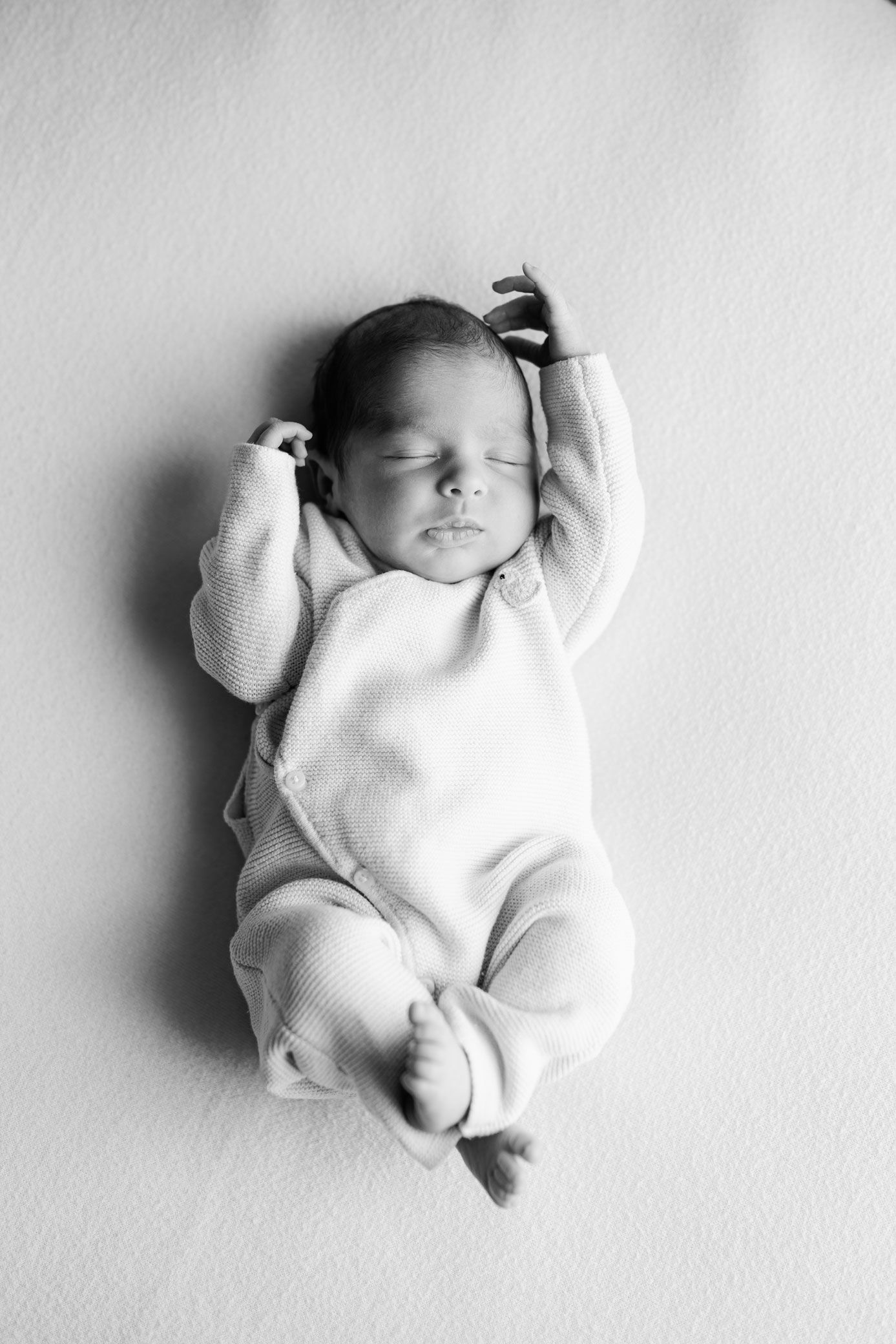 Black and white photo of a sleeping newborn baby in a cozy knit wrap outfit with both arms stretched above the head.