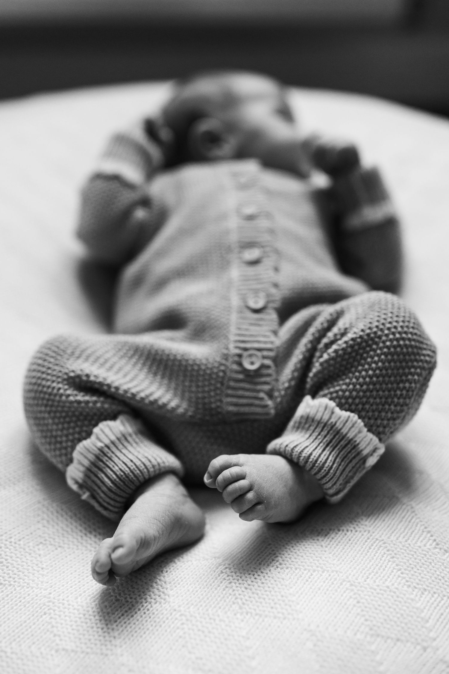 Black and white photo of a newborn baby lying on a white blanket wearing a knit romper with tiny bare feet.