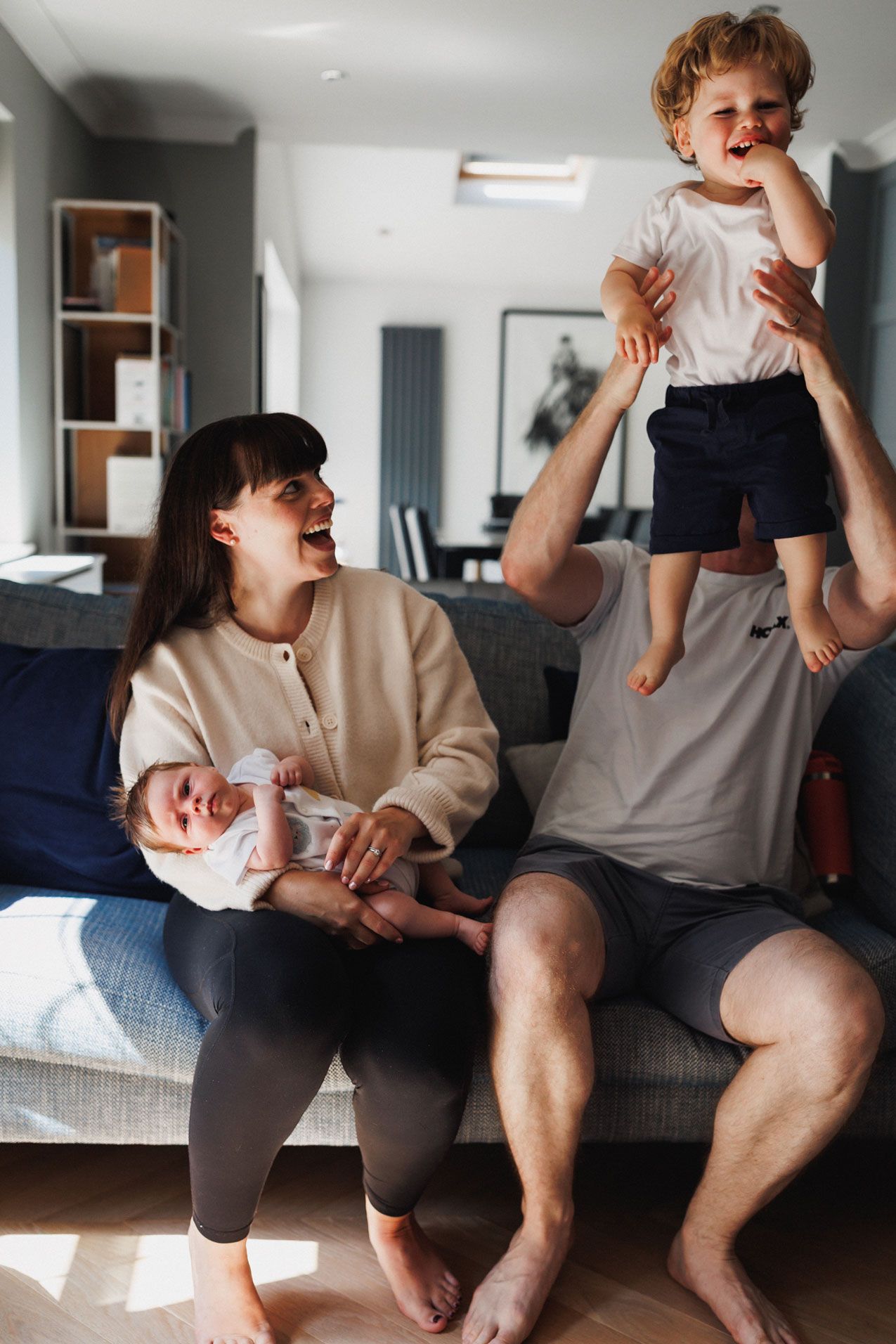 Happy family sitting on sofa, mother holding newborn baby while father lifts laughing toddler in bright living room.