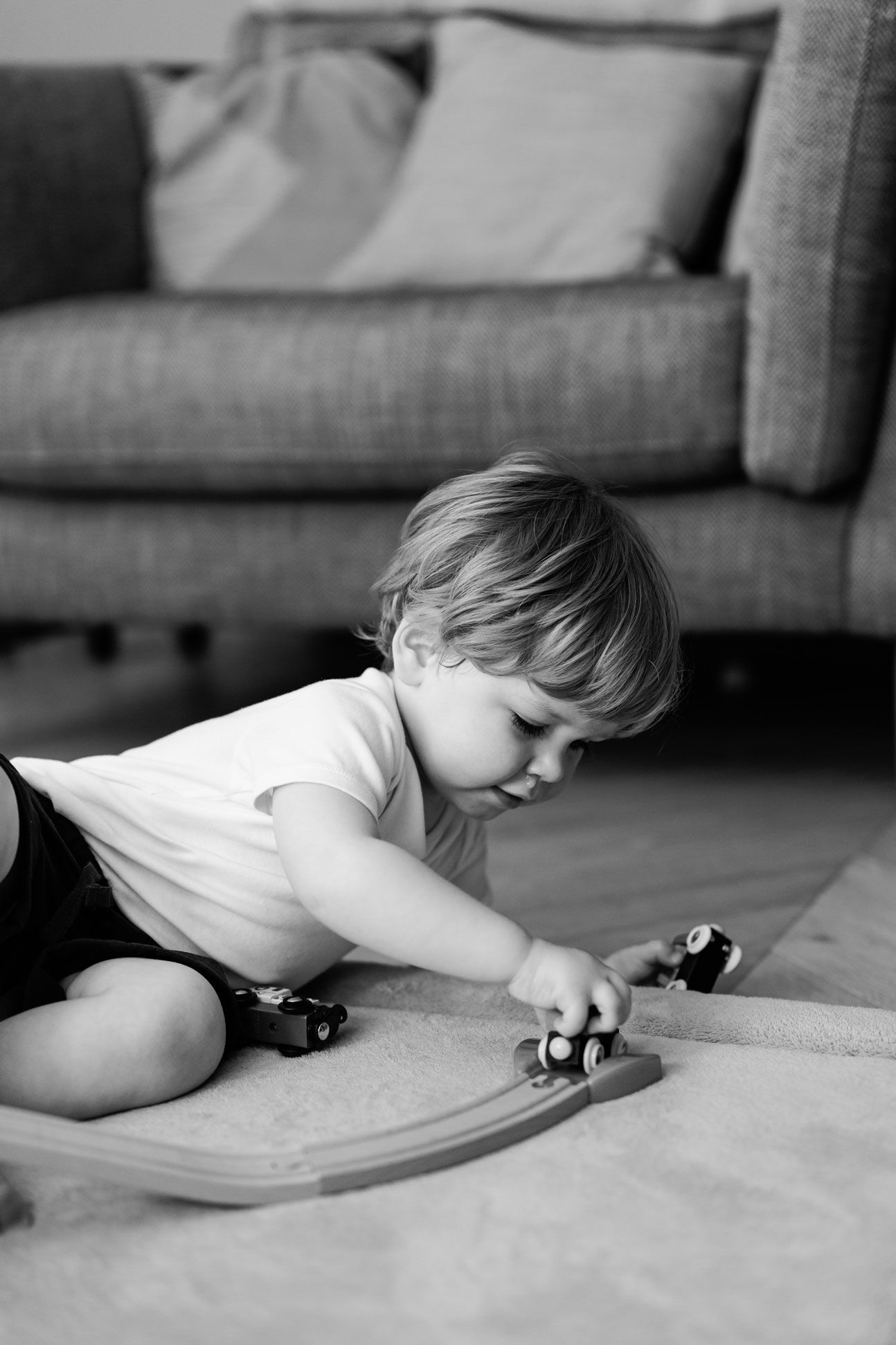 Black and white photo of a toddler boy playing with wooden toy trains on a track on the floor at home.
