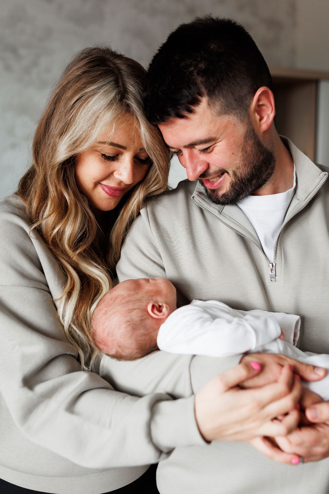 Smiling couple in grey sweatshirts tenderly holding their newborn baby close together in a nursery room.