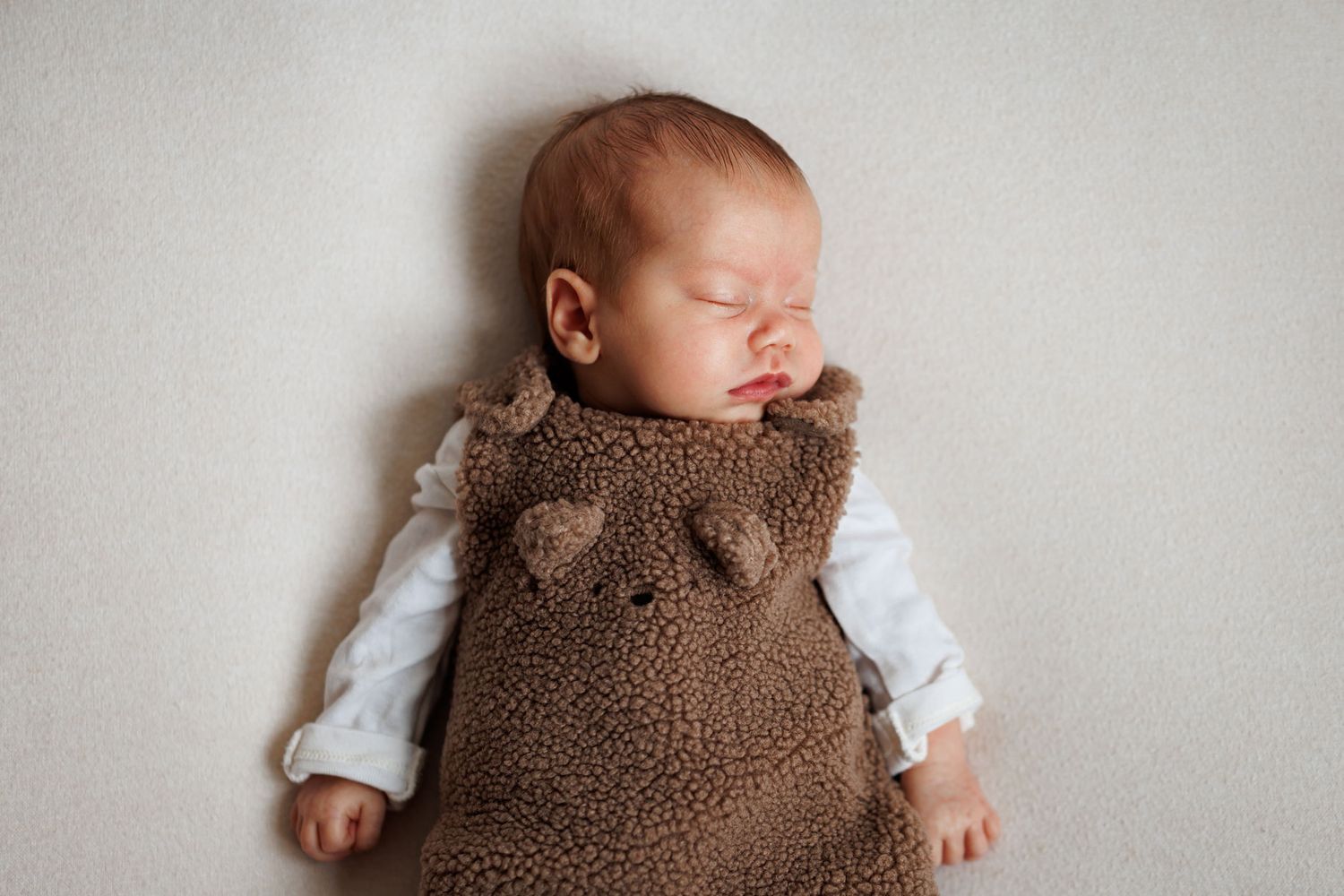 Sleeping newborn baby in a brown teddy bear fleece romper with ears over a white shirt, lying on a soft cream background.