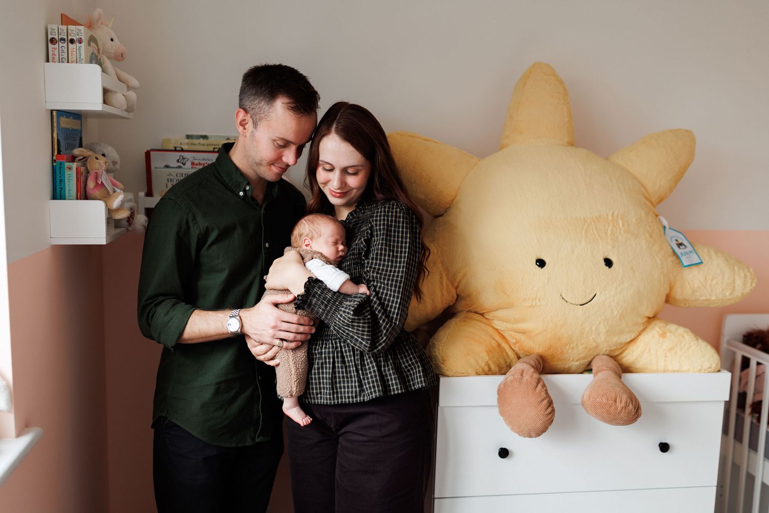 Smiling parents hold their newborn baby in a cozy nursery beside a white dresser and a large plush smiling sun pillow.