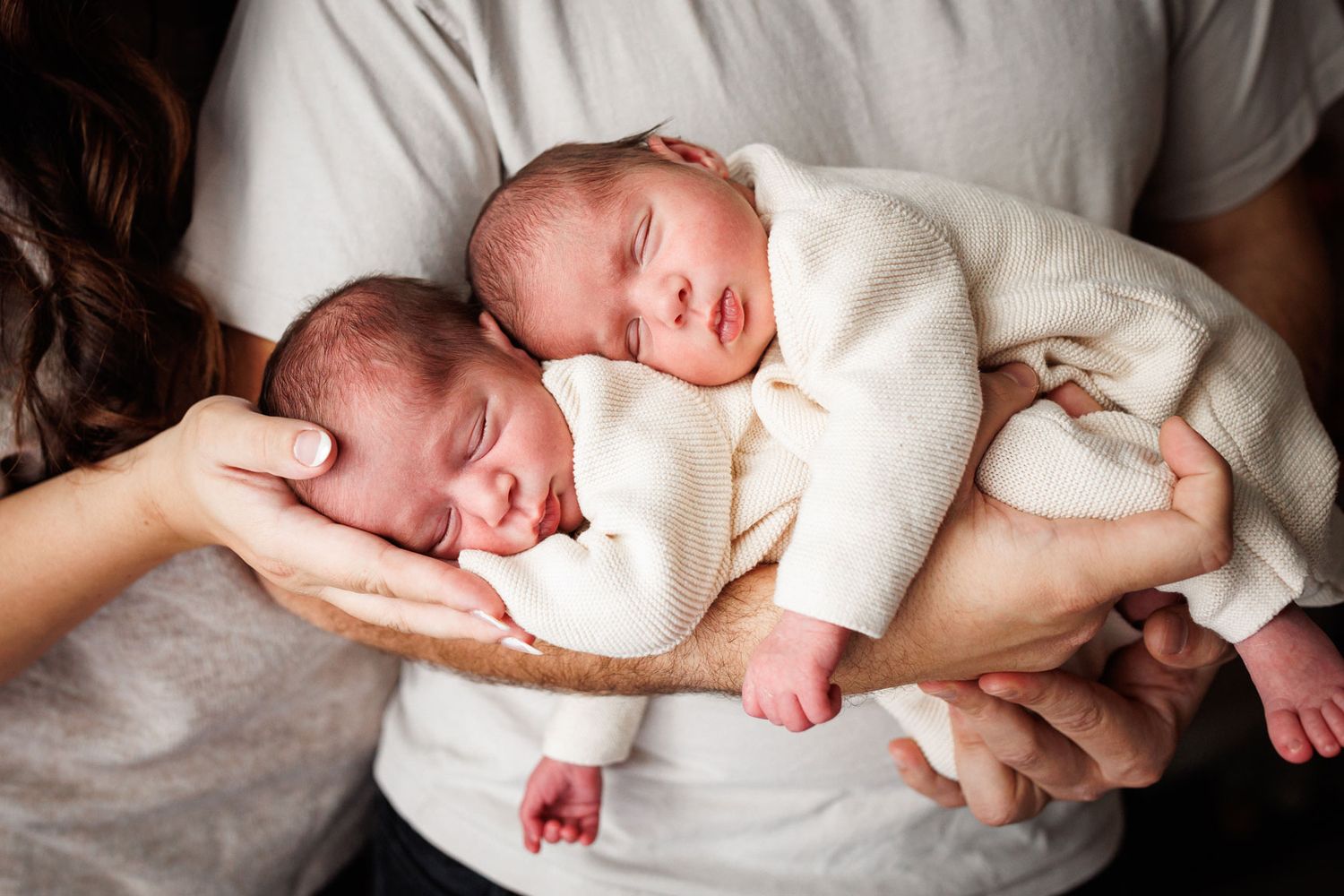 Two sleeping newborn twins in matching cream outfits rest side by side on a parent's cradled arms in a tender, peaceful family moment.