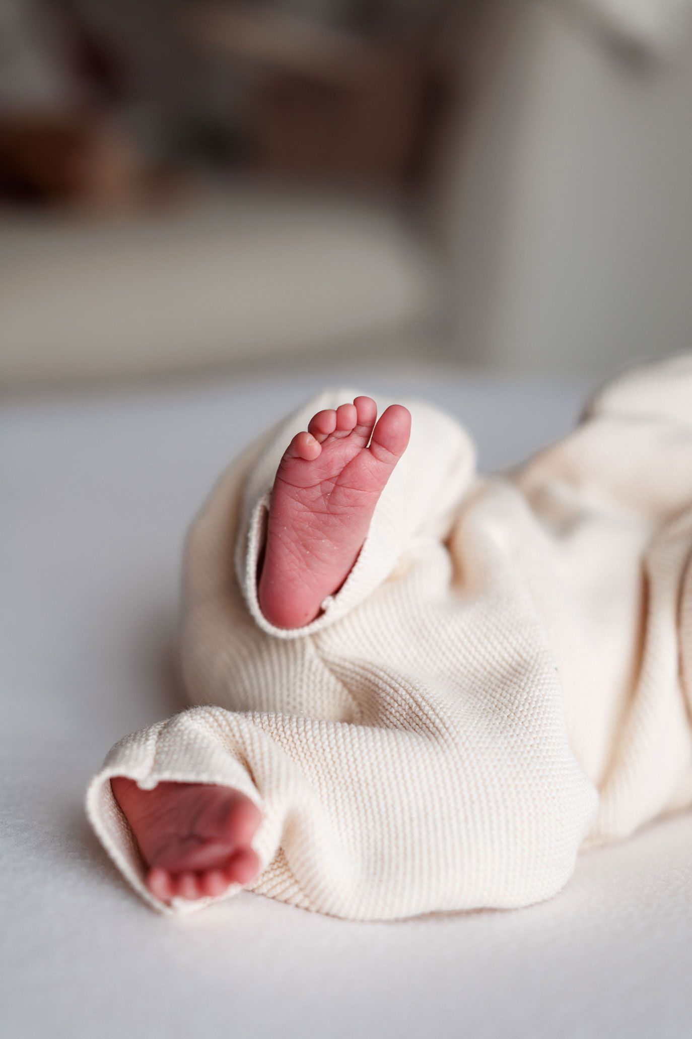 Tiny newborn baby feet with pink toes peeking out of a cream knit outfit resting on a soft white blanket.
