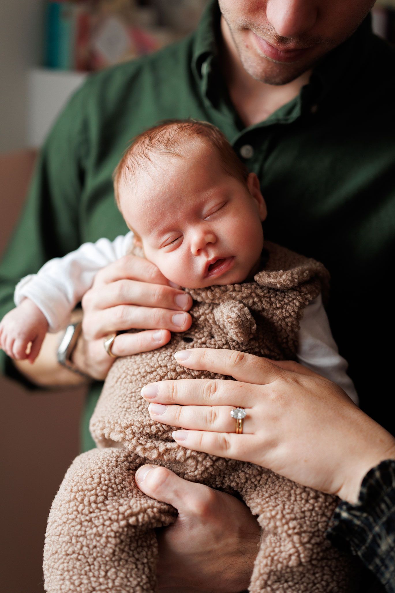 Sleeping newborn baby in a brown teddy bear fleece outfit being cradled by two parents' hands, one wearing a diamond engagement ring.